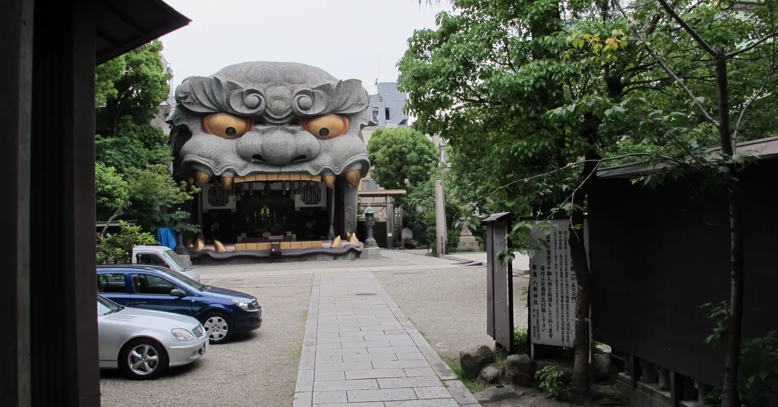The lion-head stage at Namba Yasaka Shrine in Osaka, a grey sculpted lion's face two storeys high with gold eyes and an open mouth forming the stage beneath, seen down a paved shrine path between trees