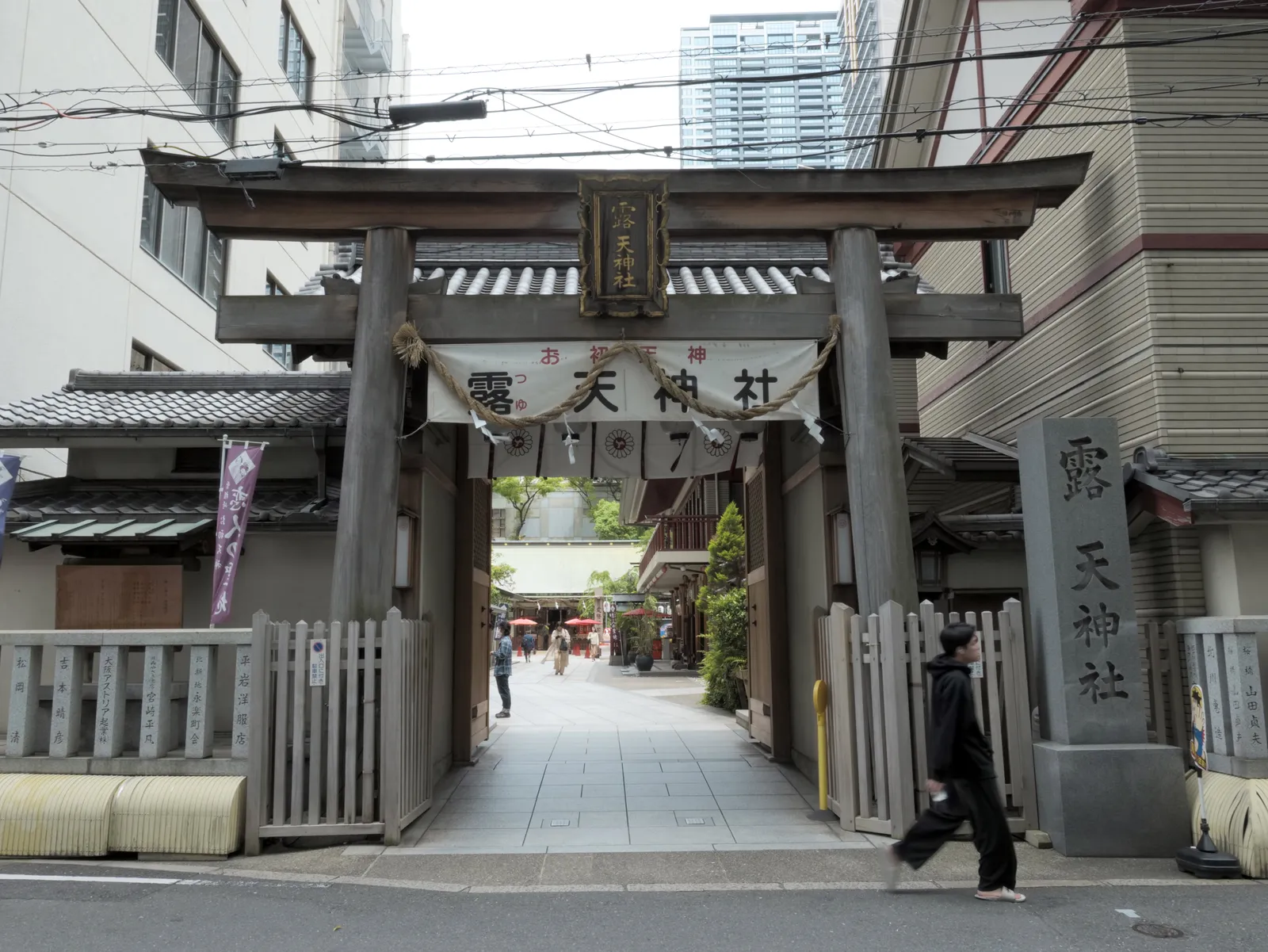 The timber torii gate of Tsuyunoten Shrine in Osaka, hung with a straw rope and a wooden name board, standing in a narrow street with apartment and office blocks pressing in on both sides