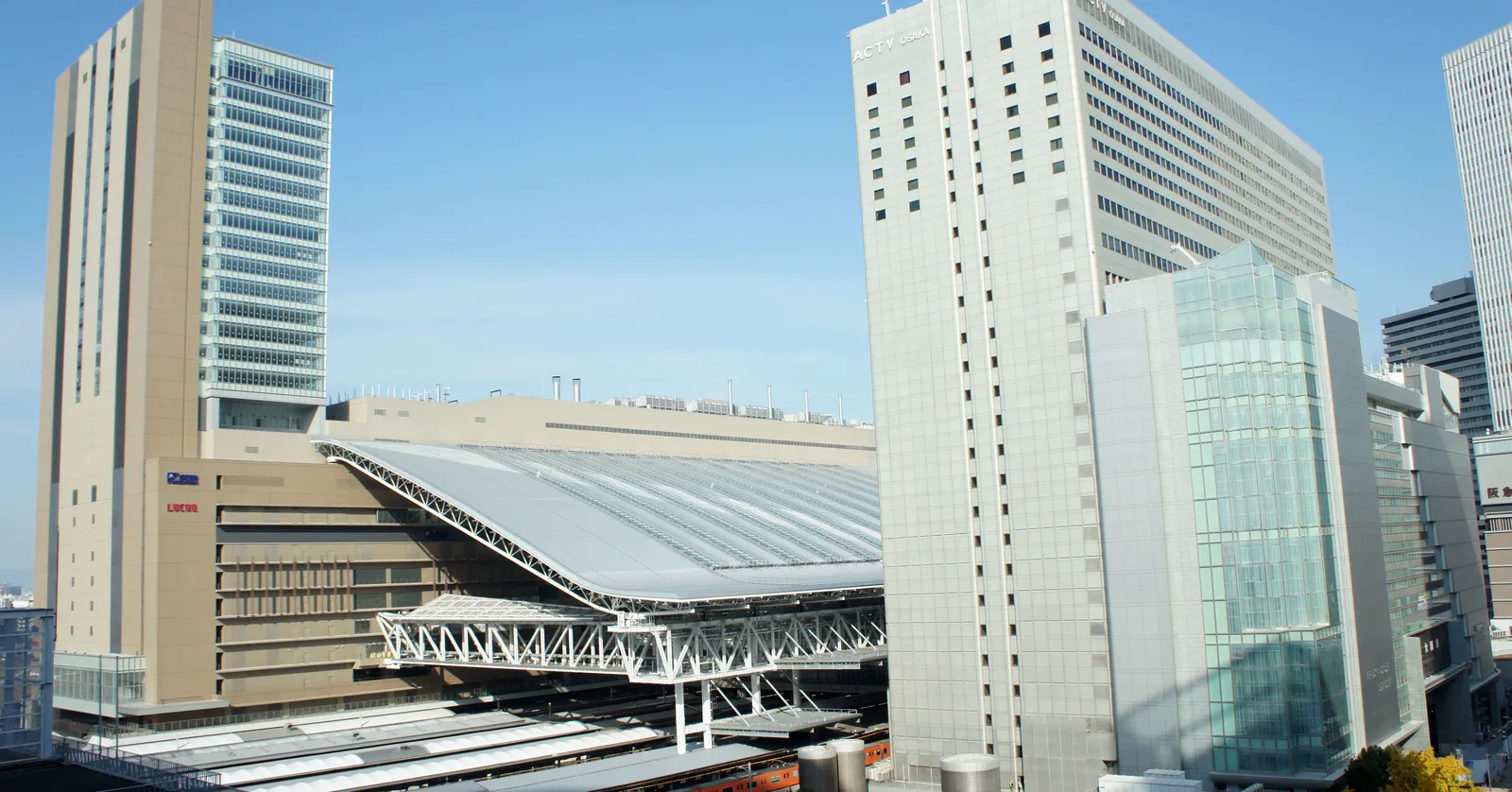 Osaka Station seen from across the tracks, a wide curved glass and steel canopy spanning the platforms with the tall pale North Gate Building and other office towers standing around it against a clear sky