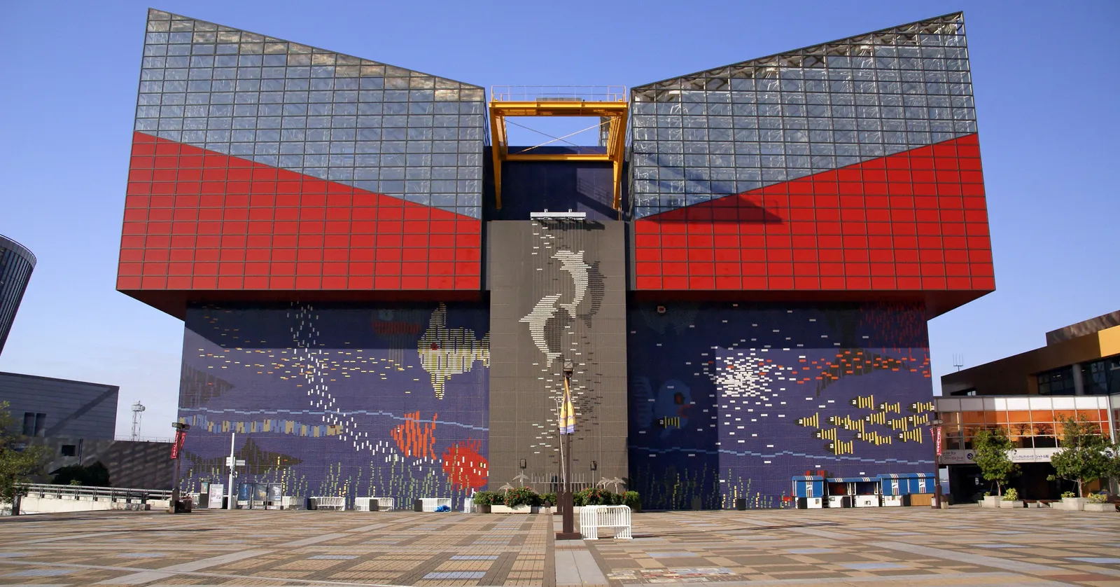 The front of Osaka Aquarium Kaiyukan, a block of red, blue and glass panels above a wide ground-floor mural of fish and sea creatures in mosaic tile, with an empty paved plaza in front of it