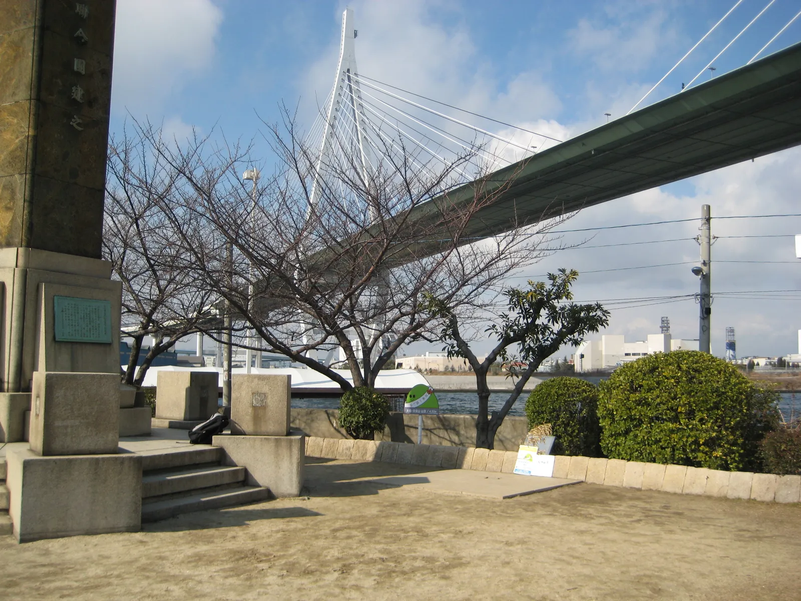 Tempozan Park on the Osaka waterfront, a stone monument and low steps under bare trees with the green cable-stayed deck of the Tempozan Bridge crossing overhead and the harbour water beyond