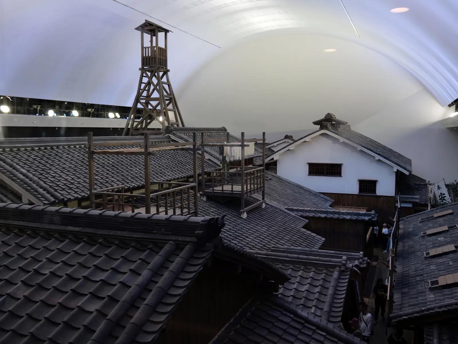 The reconstructed Edo-period townscape inside the Osaka Museum of Housing and Living, a block of full-size tiled roofs and a timber fire lookout tower with a white plastered storehouse beyond, all standing under a curved indoor ceiling