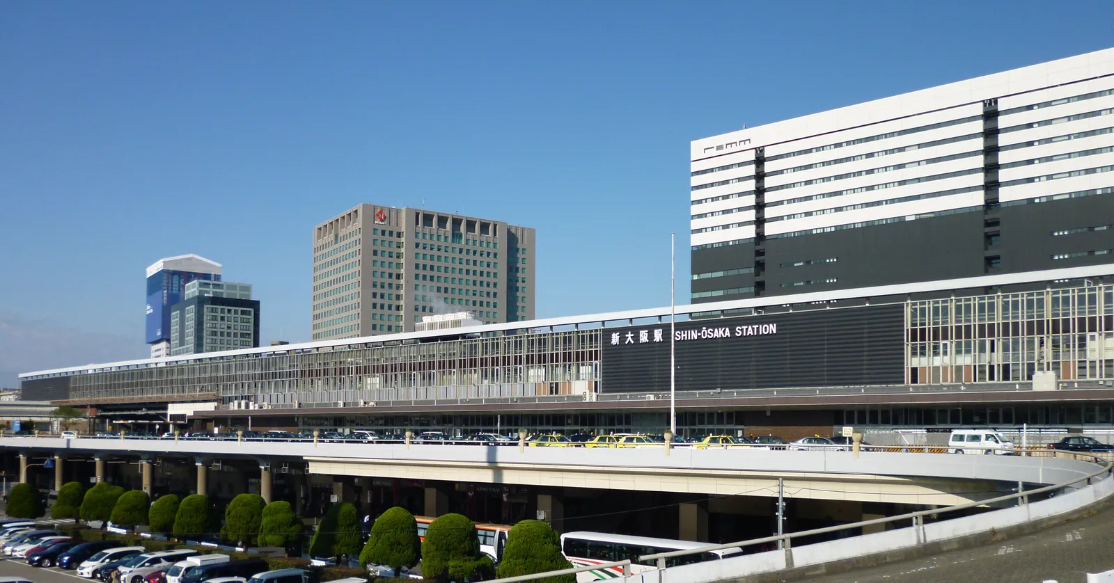 Shin-Osaka station from the street, a long elevated concrete structure with SHIN-OSAKA STATION lettered along its front, viaducts running out of both ends and office towers standing behind it