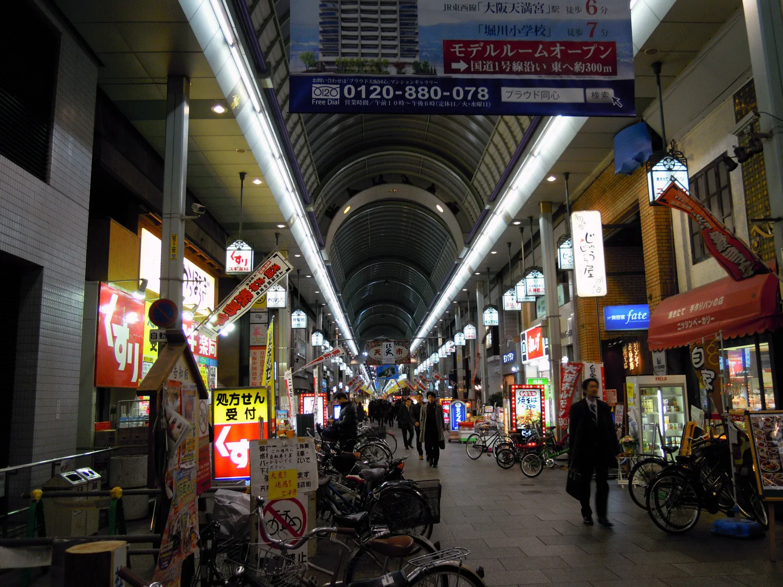 The Tenjinbashisuji arcade in Osaka running away into the distance under a long curved roof, shopfronts and hanging signboards down both sides and rows of parked bicycles along the edges