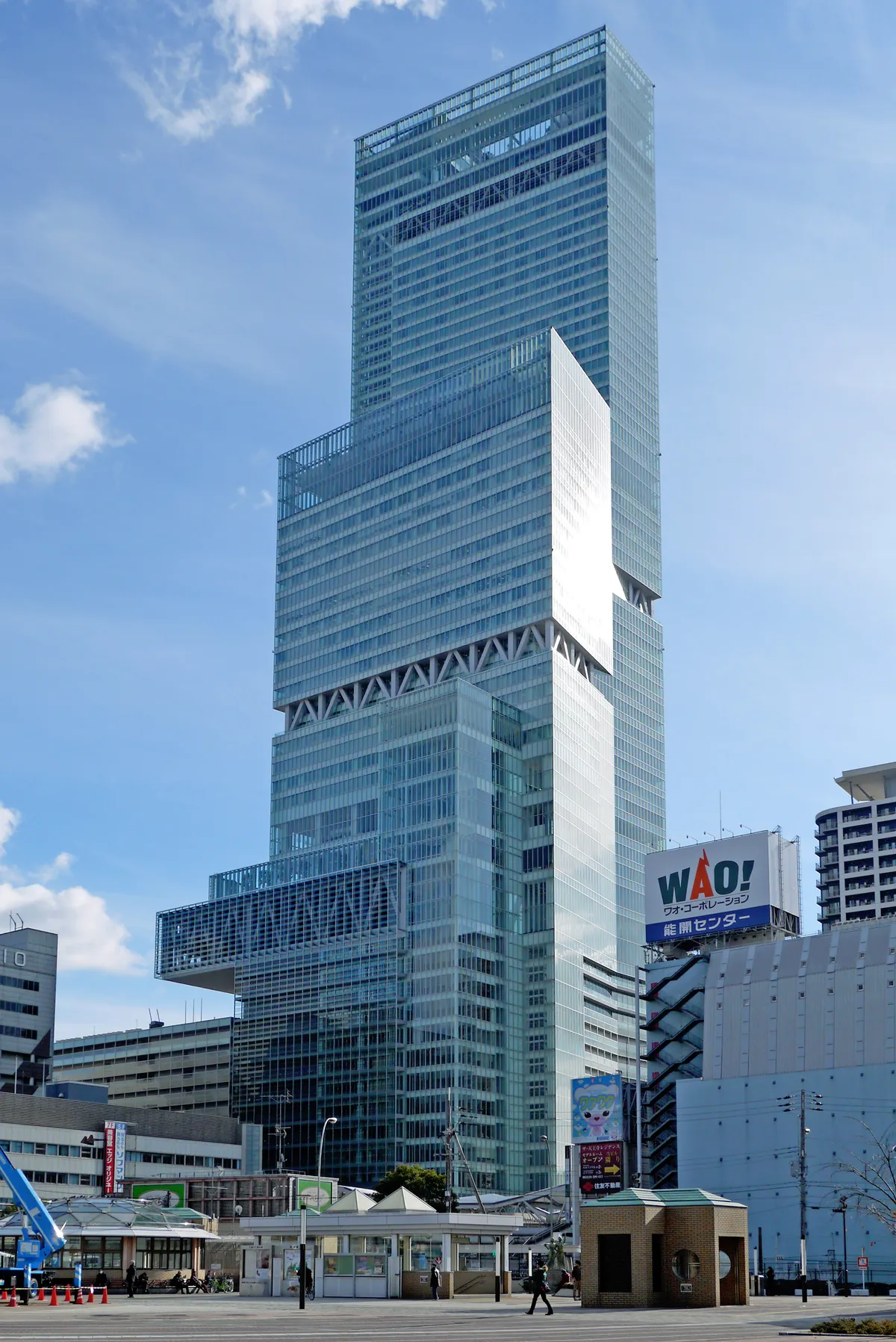 The upper floors of Abeno Harukas in Osaka, a stepped glass tower with three setbacks in its facade, rising against a blue sky above lower buildings