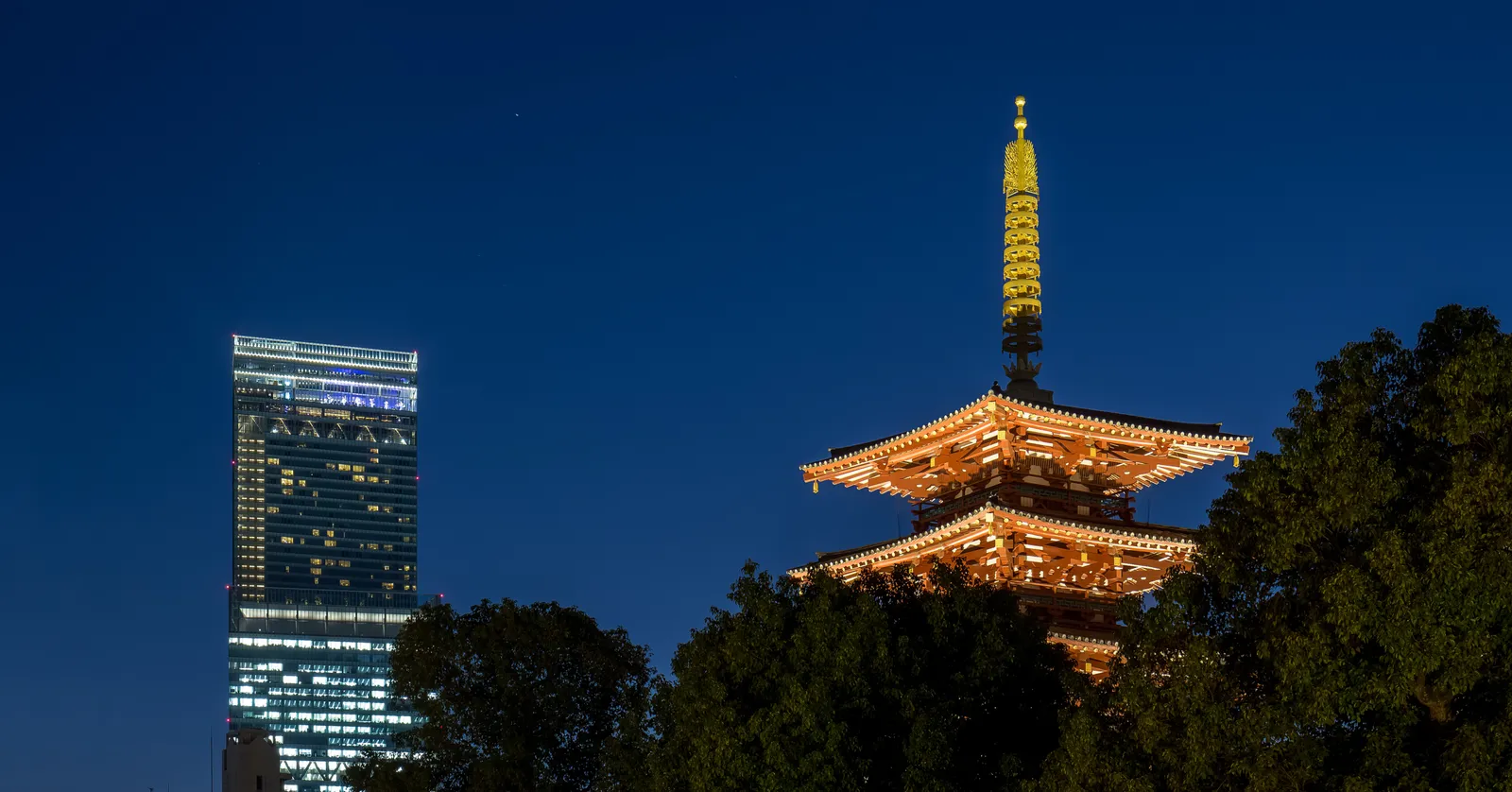 The illuminated five-storey pagoda of Shitennoji lit orange against a deep blue dusk sky, with the lit glass slab of the Abeno Harukas tower standing behind it to the left above dark trees