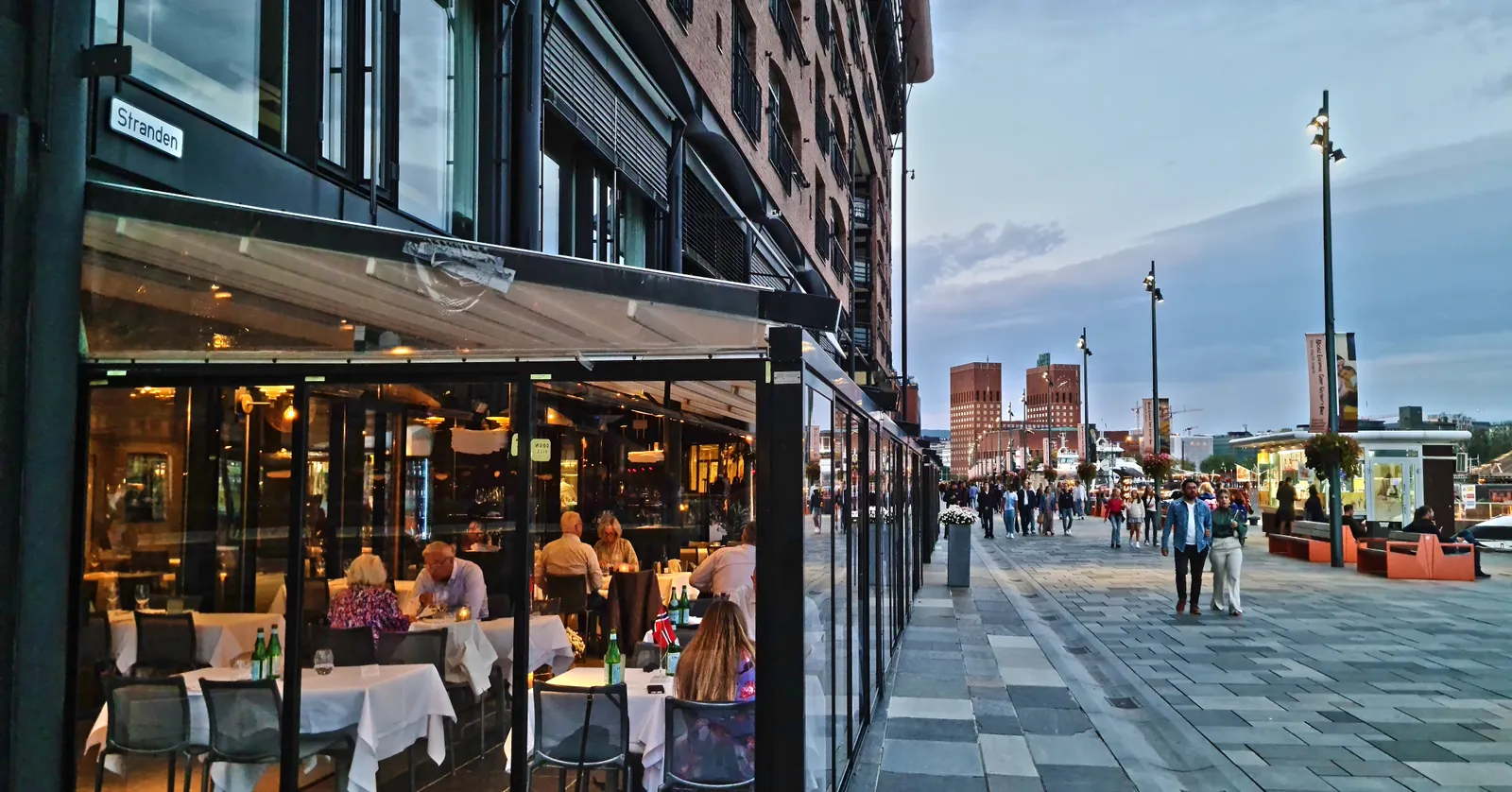 The Aker Brygge boardwalk in Oslo at dusk, glass-fronted restaurant terraces along a brick warehouse frontage on the left and people walking the wide paved quay towards the harbour