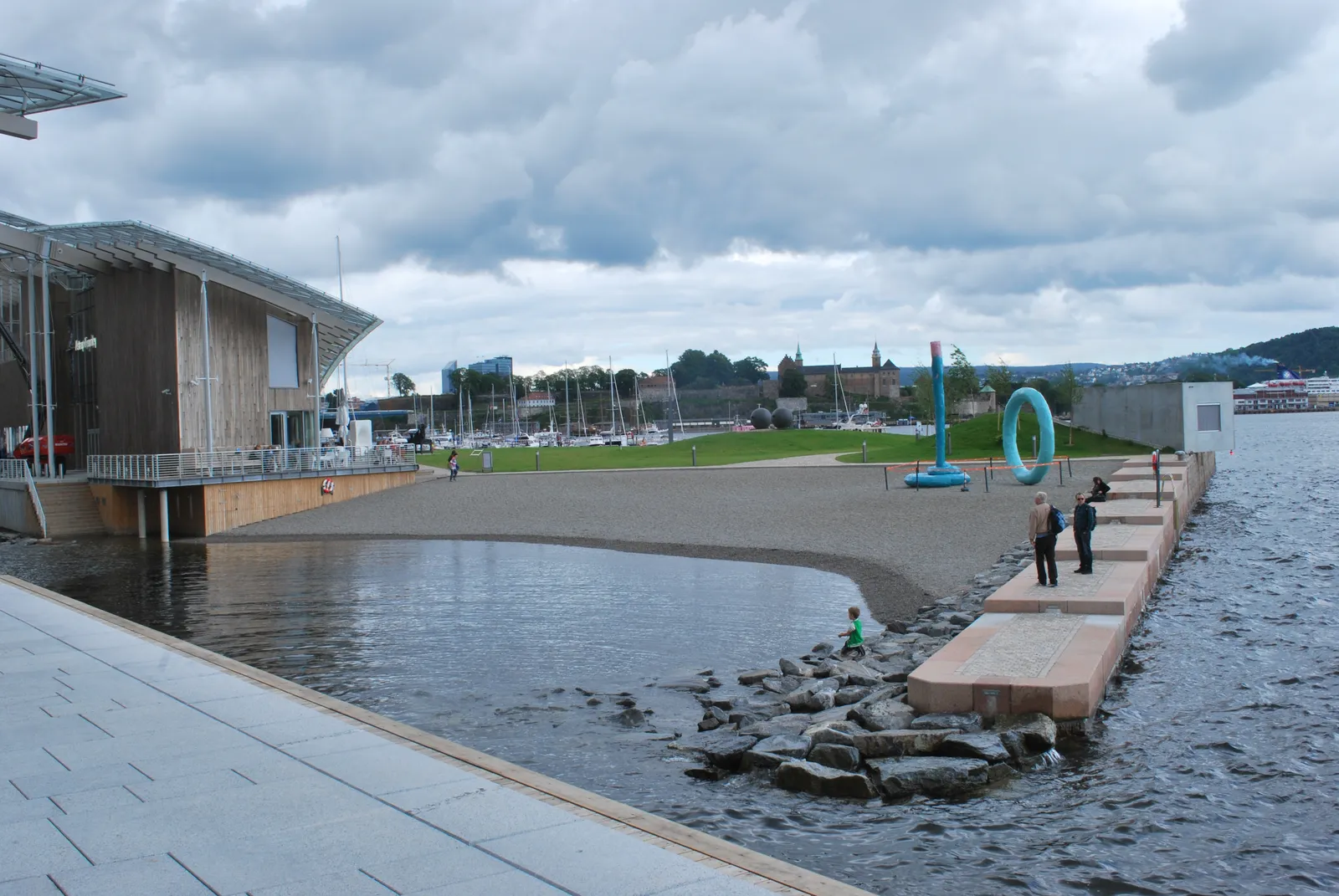 The small sand beach at the tip of Tjuvholmen in Oslo, the timber wall and glass roof of the art museum on the left, the fjord lapping against a stone edge and people standing along the quay