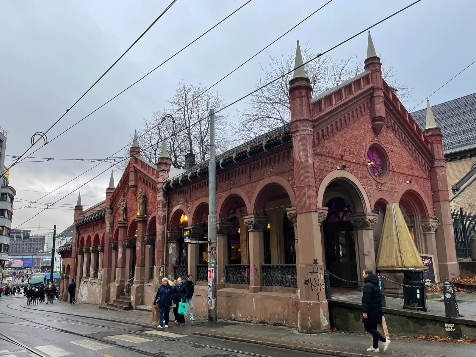 The Basarhallene market arcade behind Oslo Cathedral, a long red brick building of open round arches with small pointed turrets along its roofline, tram wires strung across the street in front