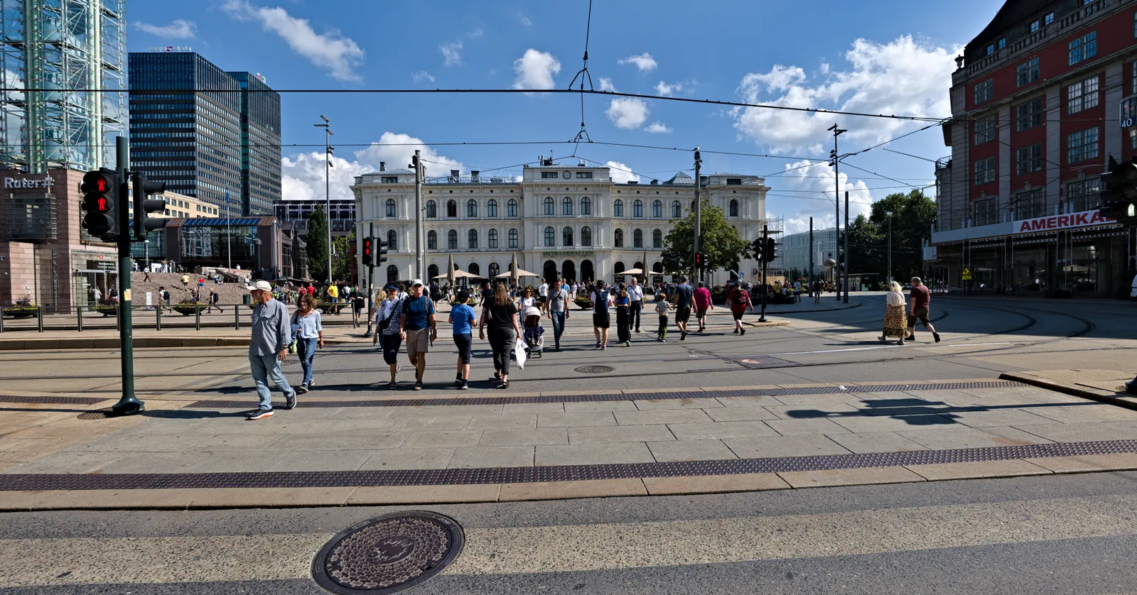 Jernbanetorget square outside Oslo Central Station on a summer day, tram wires overhead and people crossing the paving, with the white 19th-century Østbanehallen building standing across the square
