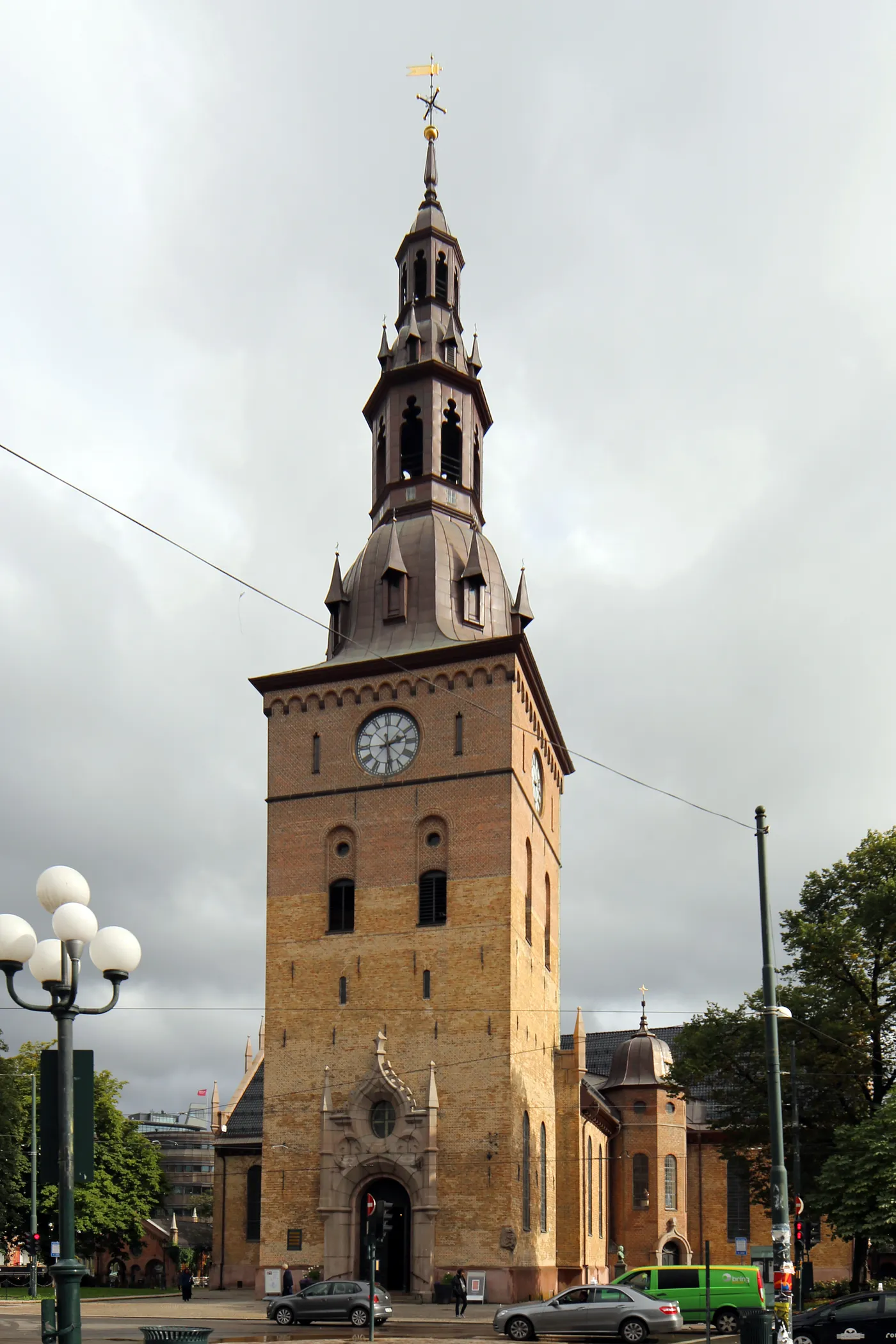 Oslo Cathedral seen from the street, a yellow-brown brick church with a clock face on its tower and a dark copper spire and bell stage above it