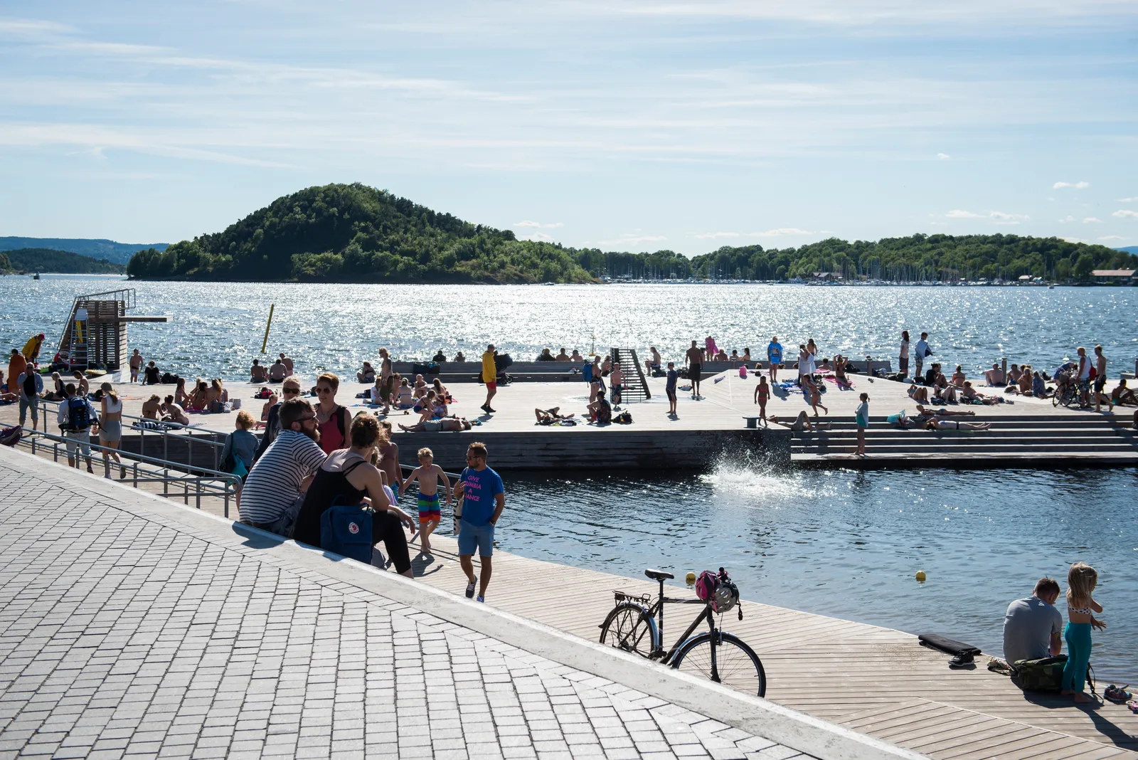 Sørenga seawater pool in Oslo on a sunny day, people sitting along the stone steps and out on the floating wooden jetties in the fjord, with a wooded island rising behind