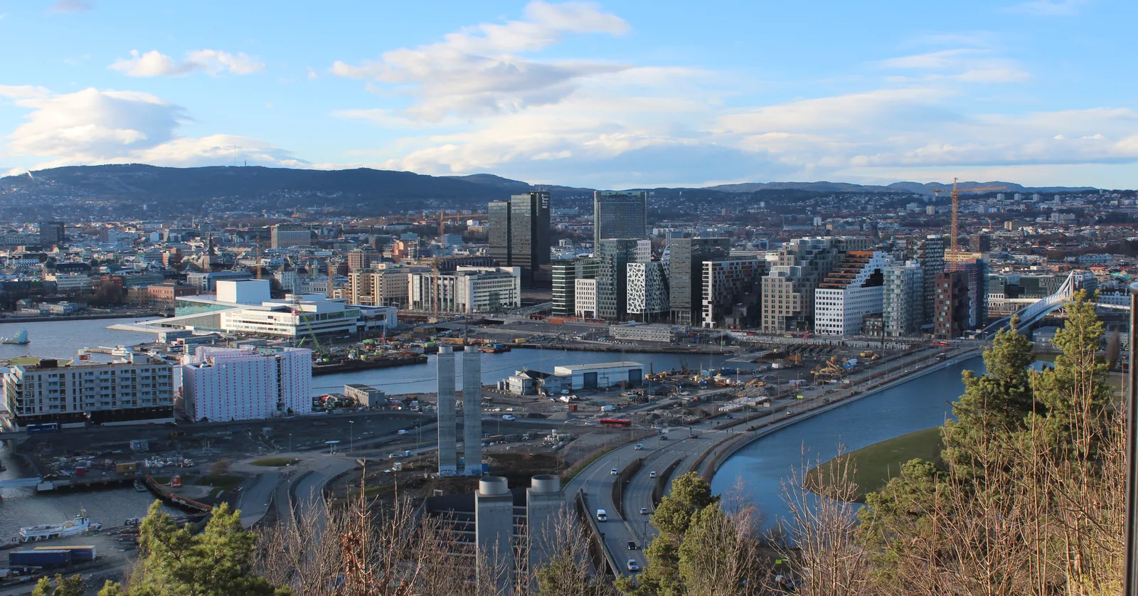 Oslo seen from the Ekeberg ridge, the harbour and the row of tall waterfront blocks in the middle distance, a river and rail lines below, and wooded hills closing the far side of the city