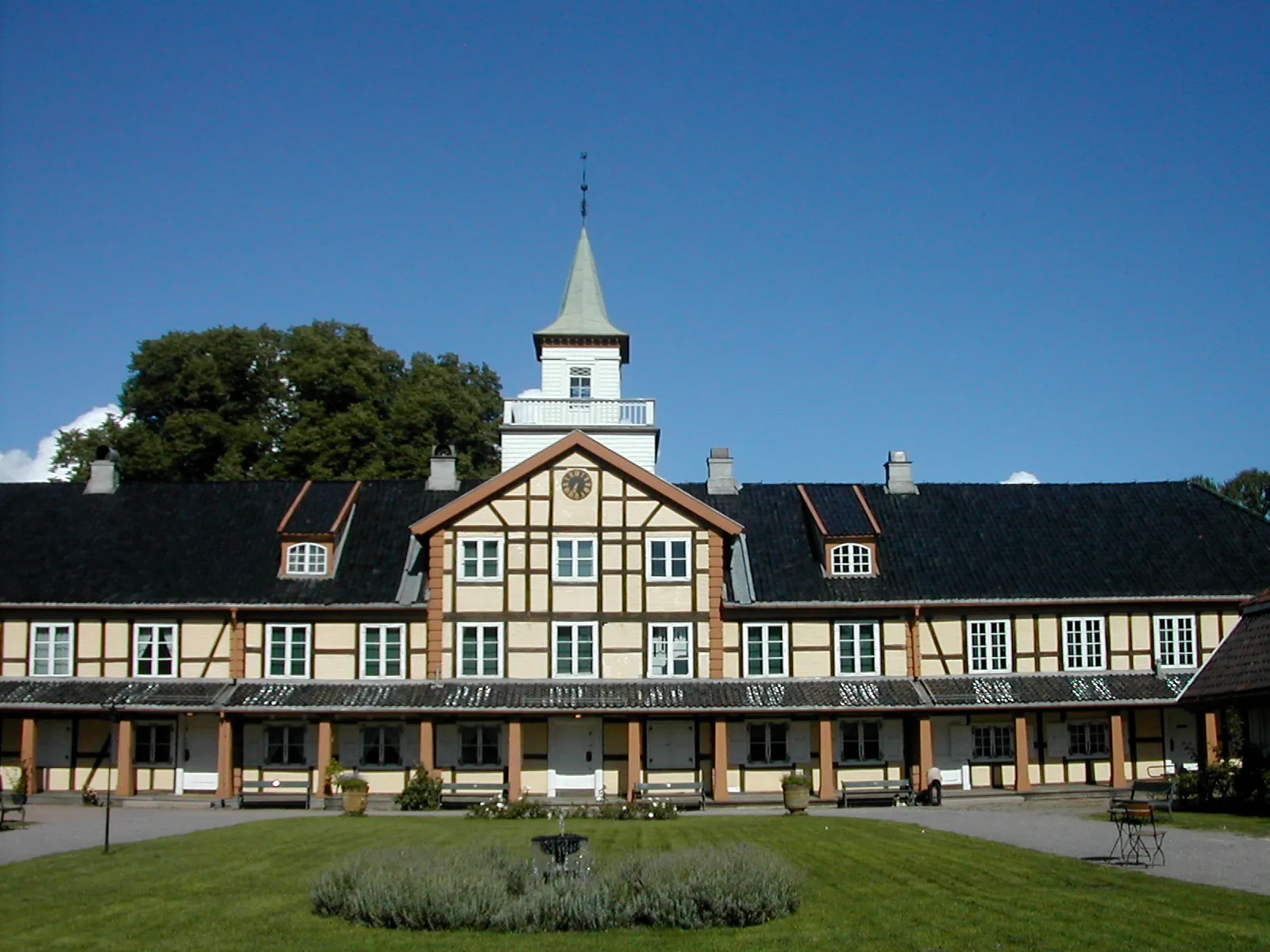 Frogner Manor in Oslo on a clear summer day, a long half-timbered range around a courtyard with a covered arcade at ground level and a white clock tower rising from the roof