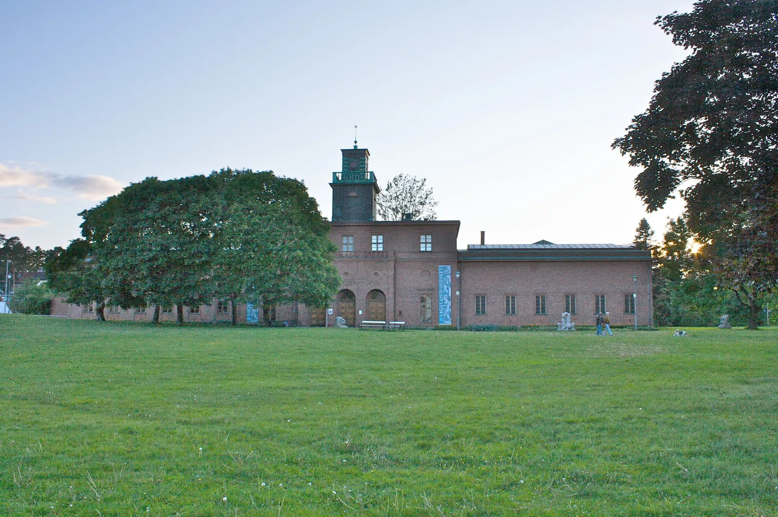 The Vigeland Museum in Oslo seen across a lawn at dusk, a long low brick building with a square tower and a green copper roof at one end