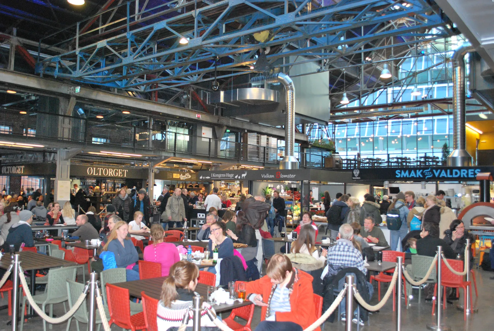 Inside Mathallen food hall in Oslo, rows of red chairs and tables filled with people under an industrial roof of exposed steel trusses, with lit food counters along the walls on both sides