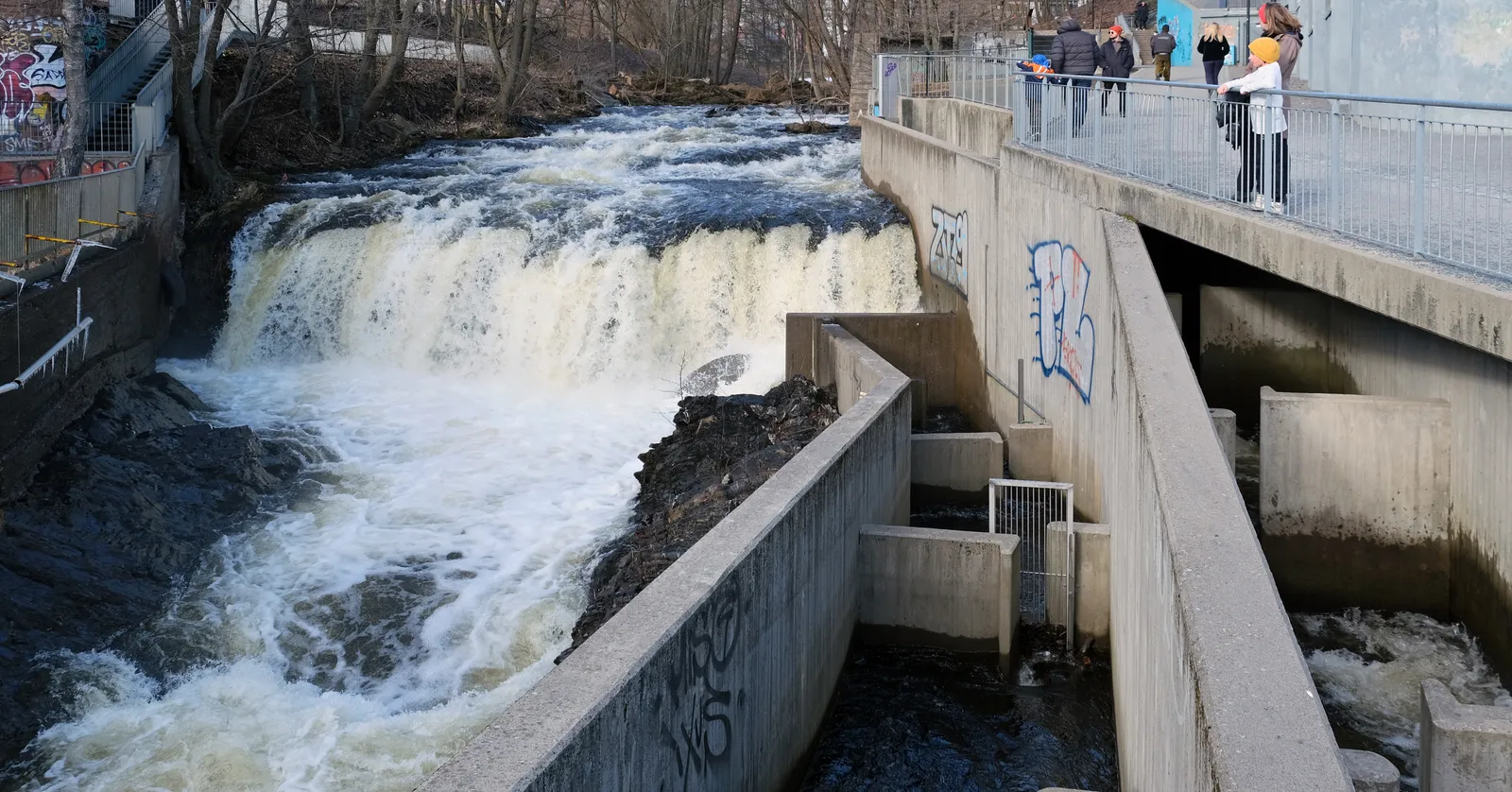 The Nedre Foss waterfall on the Akerselva in Oslo, white water dropping over a wide rock ledge beside a concrete weir, with a railed walkway and people looking down from the right