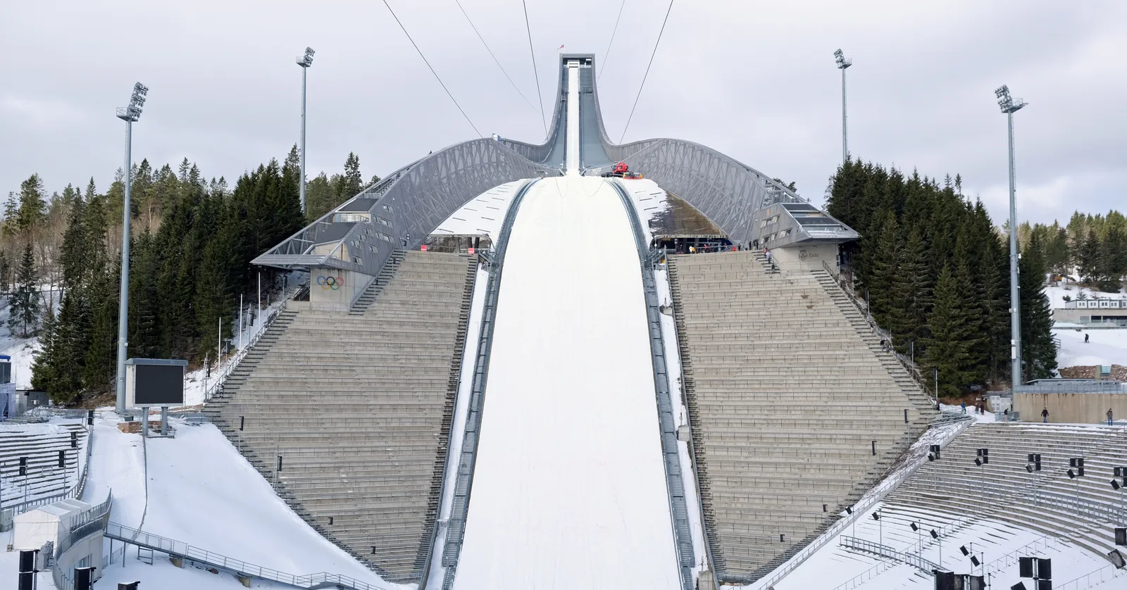 The Holmenkollen ski jump in Oslo seen head on in winter, a steel in-run running up to a cantilevered top platform, with the white landing hill below it and empty grandstands stepped up both sides