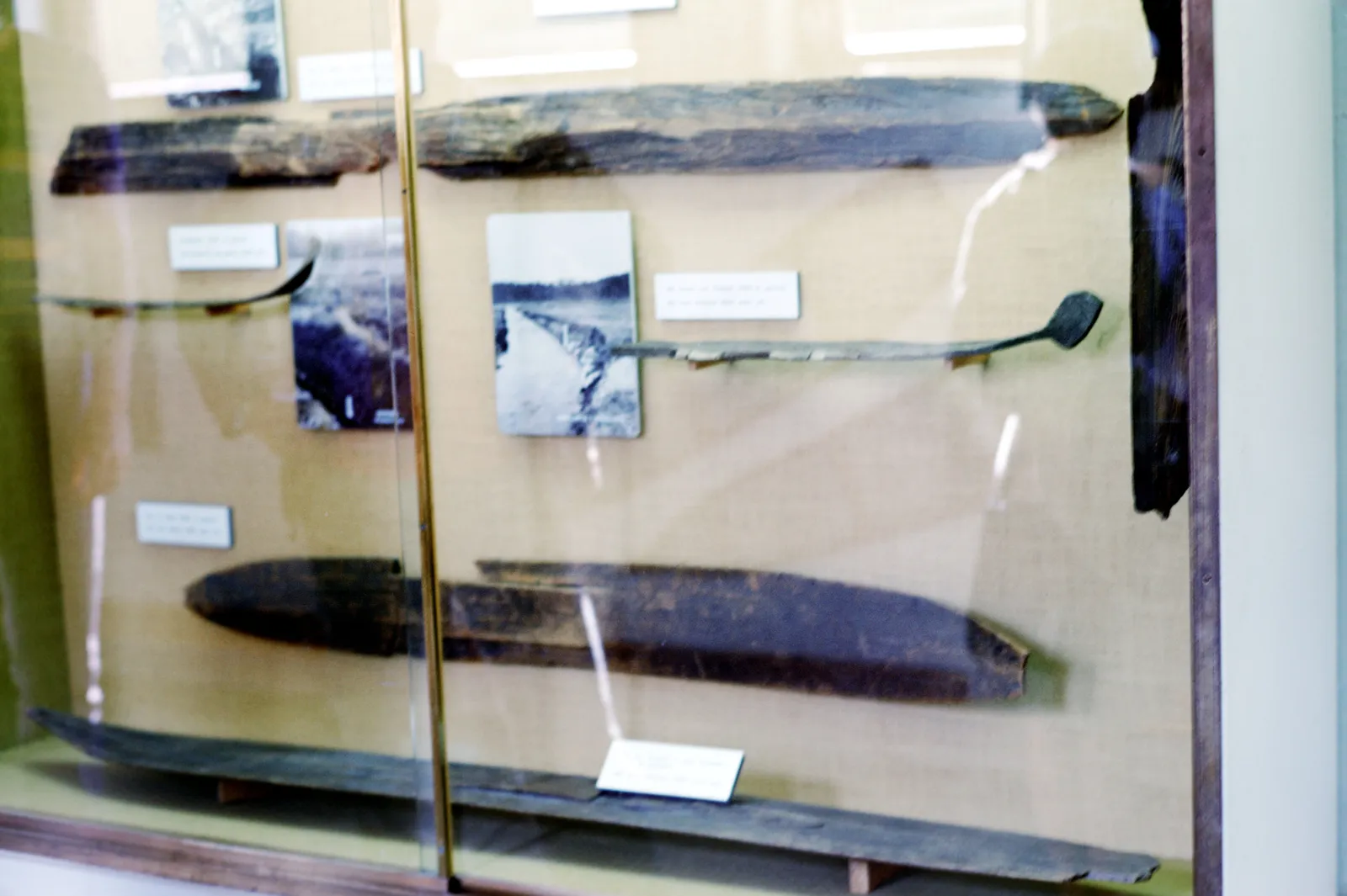 A display case in the Holmenkollen ski museum holding several dark wooden skis of different ages laid flat behind glass, with a ski pole and small printed labels beside them