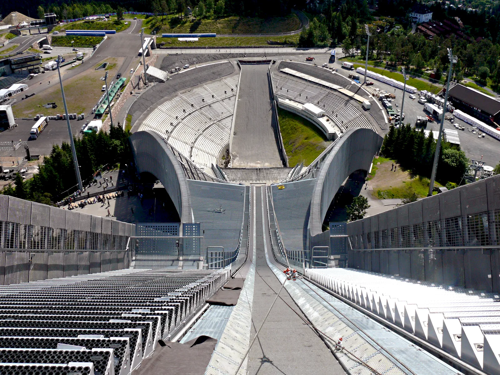 The view from the top of the Holmenkollen jump tower straight down the in-run and landing hill to the curved arena bowl at the bottom, with roads, coaches and wooded suburbs beyond