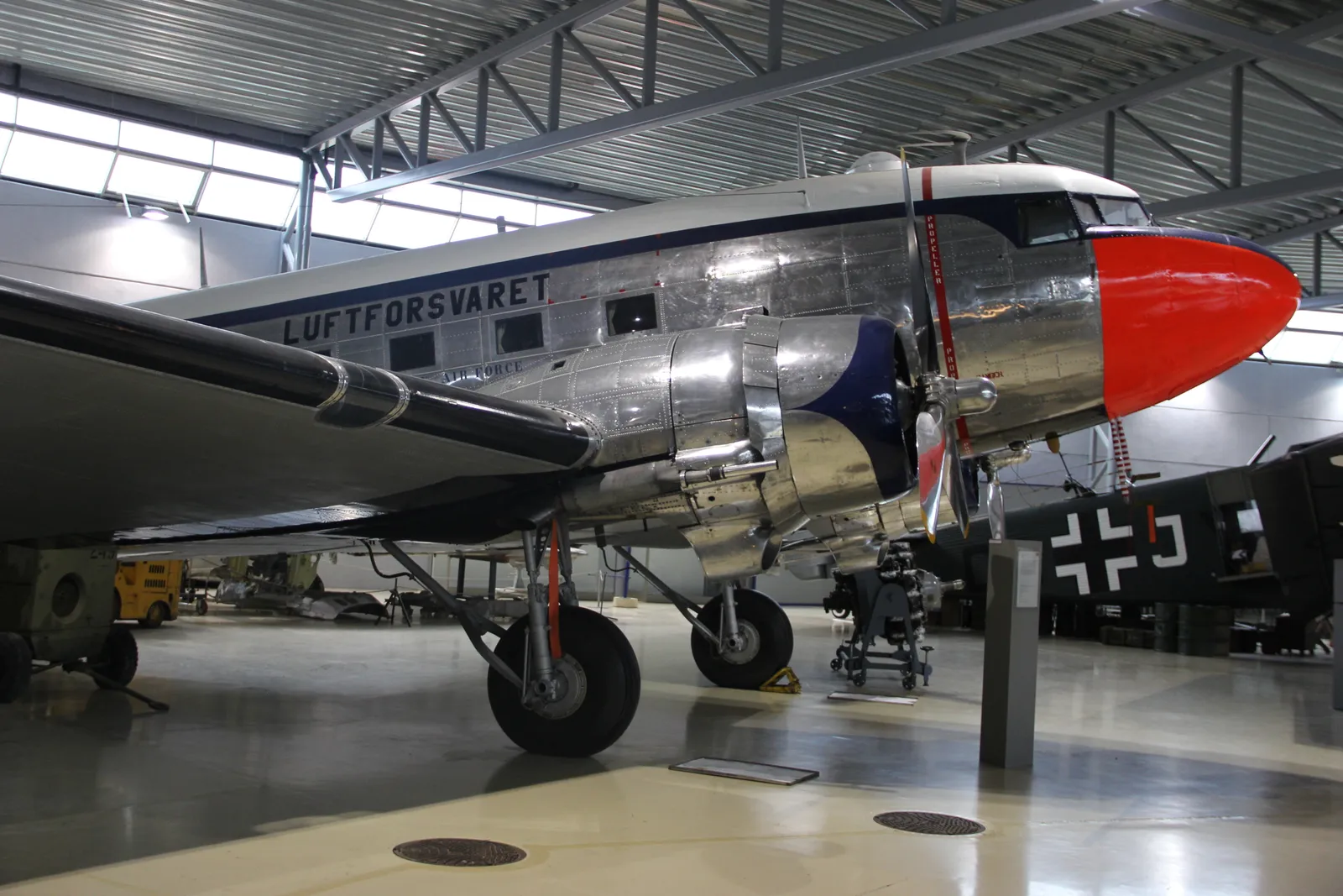 A polished silver Douglas C-47 transport aircraft lettered Luftforsvaret standing inside a museum hangar at Gardermoen, with the tails and wings of other aircraft behind it