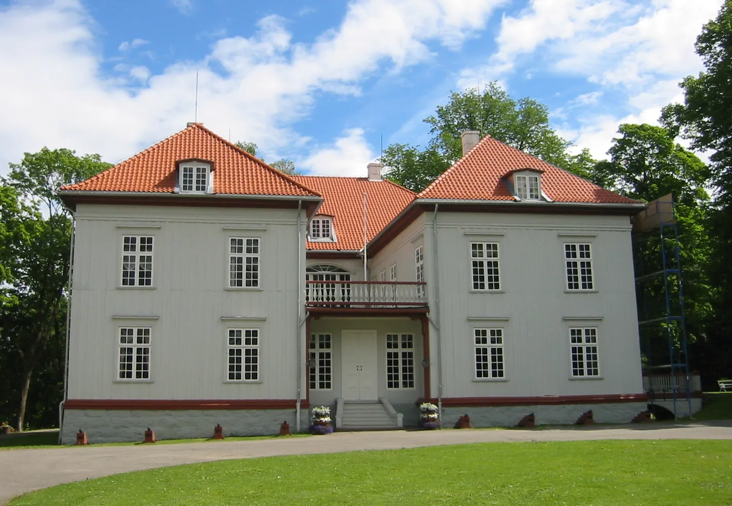 Eidsvollsbygningen, a pale grey two-storey manor house with a red tile roof and a small balcony over the front steps, standing on mown grass among trees
