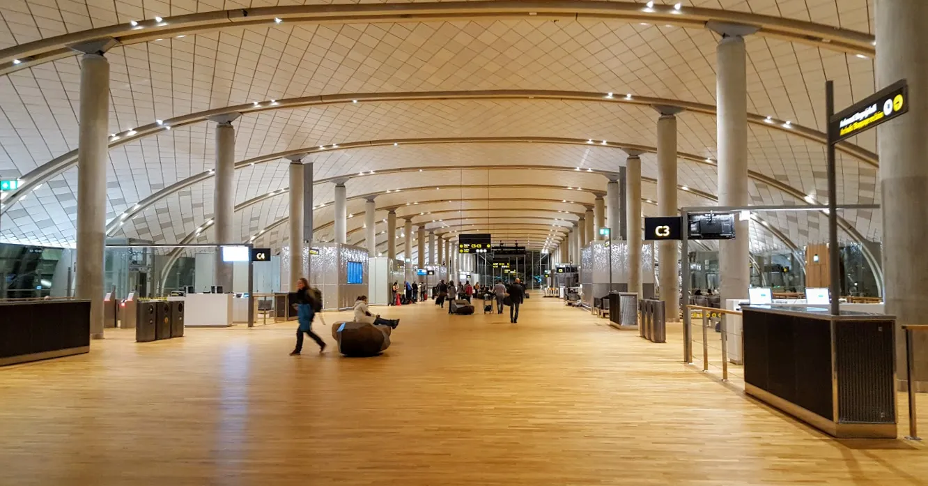 The departure pier at Oslo Airport, Gardermoen, a long hall with a pale oak floor running into the distance between rows of slim columns, under a curved roof of laminated timber beams