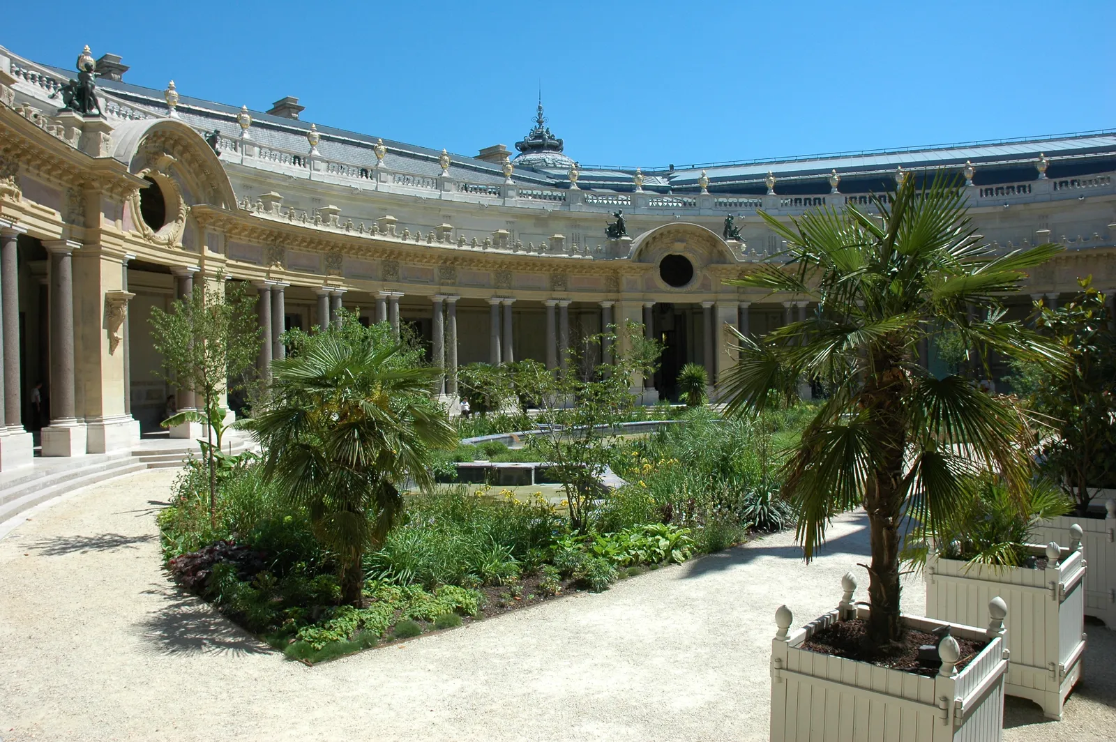 The semicircular inner garden of the Petit Palais in Paris, a colonnaded gallery curving around beds of palms and grasses with a gravel path and a mosaic-lined pool