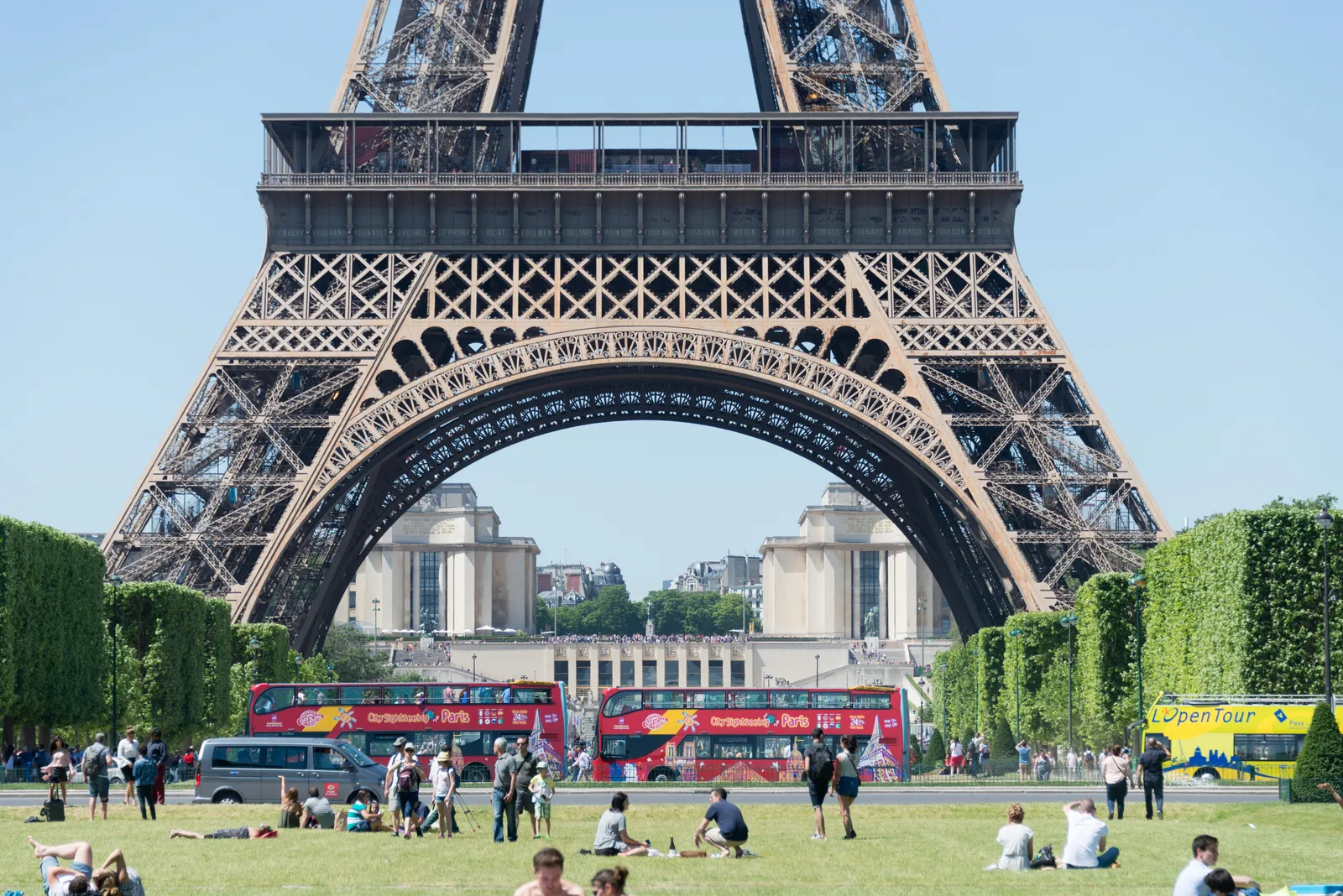 The base and first arch of the Eiffel Tower seen across the lawns of the Champ de Mars in Paris, with people sitting on the grass in the foreground