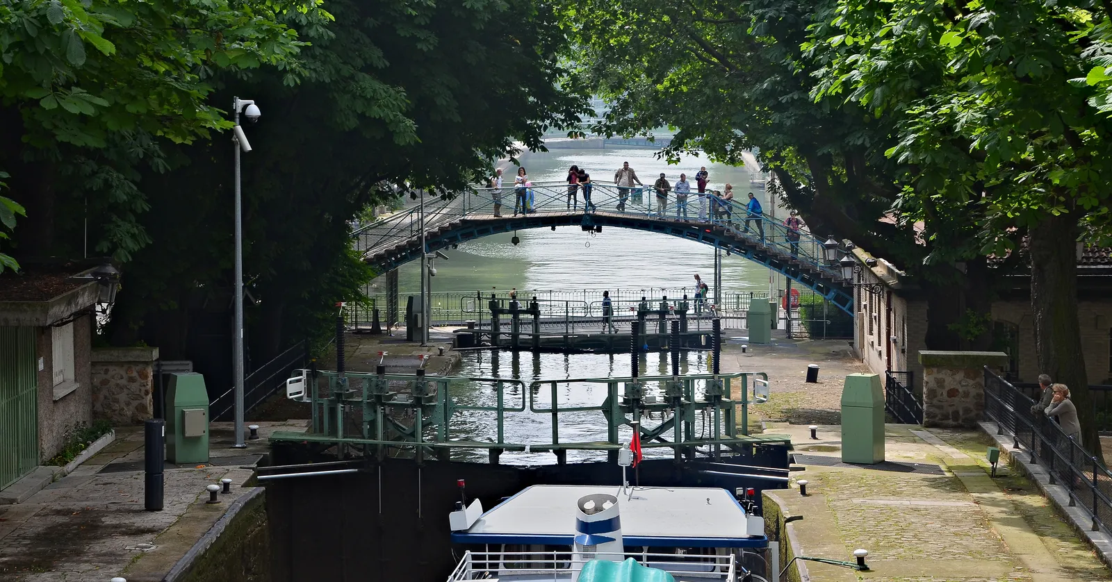 A lock on the Canal Saint-Martin in Paris with a boat waiting inside it, an arched green iron footbridge crossing above, and plane trees along both cobbled quays