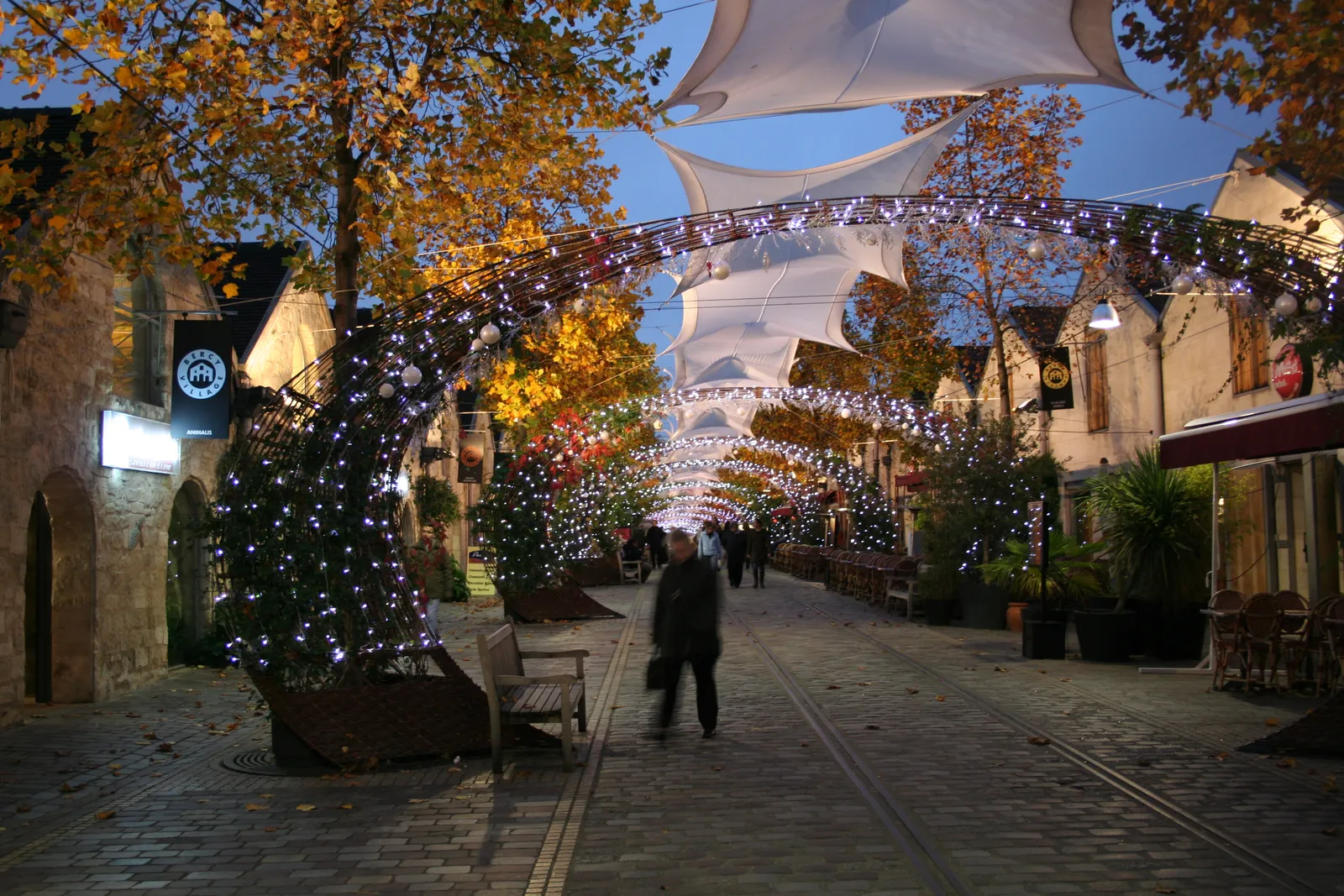 The illuminated, string-light-covered lane of Bercy Village at dusk, lined with converted stone wine warehouses now used as shops and restaurants