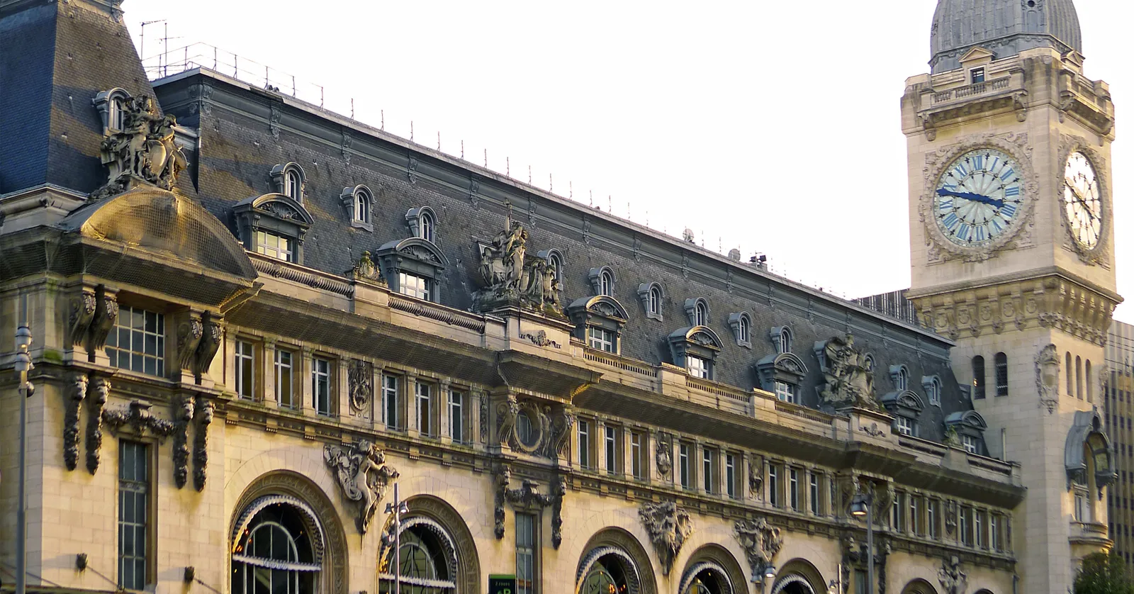 The stone facade of Paris Gare de Lyon with its arched windows and carved roofline, and the belfry clock tower with its blue-faced dial standing at the end of it