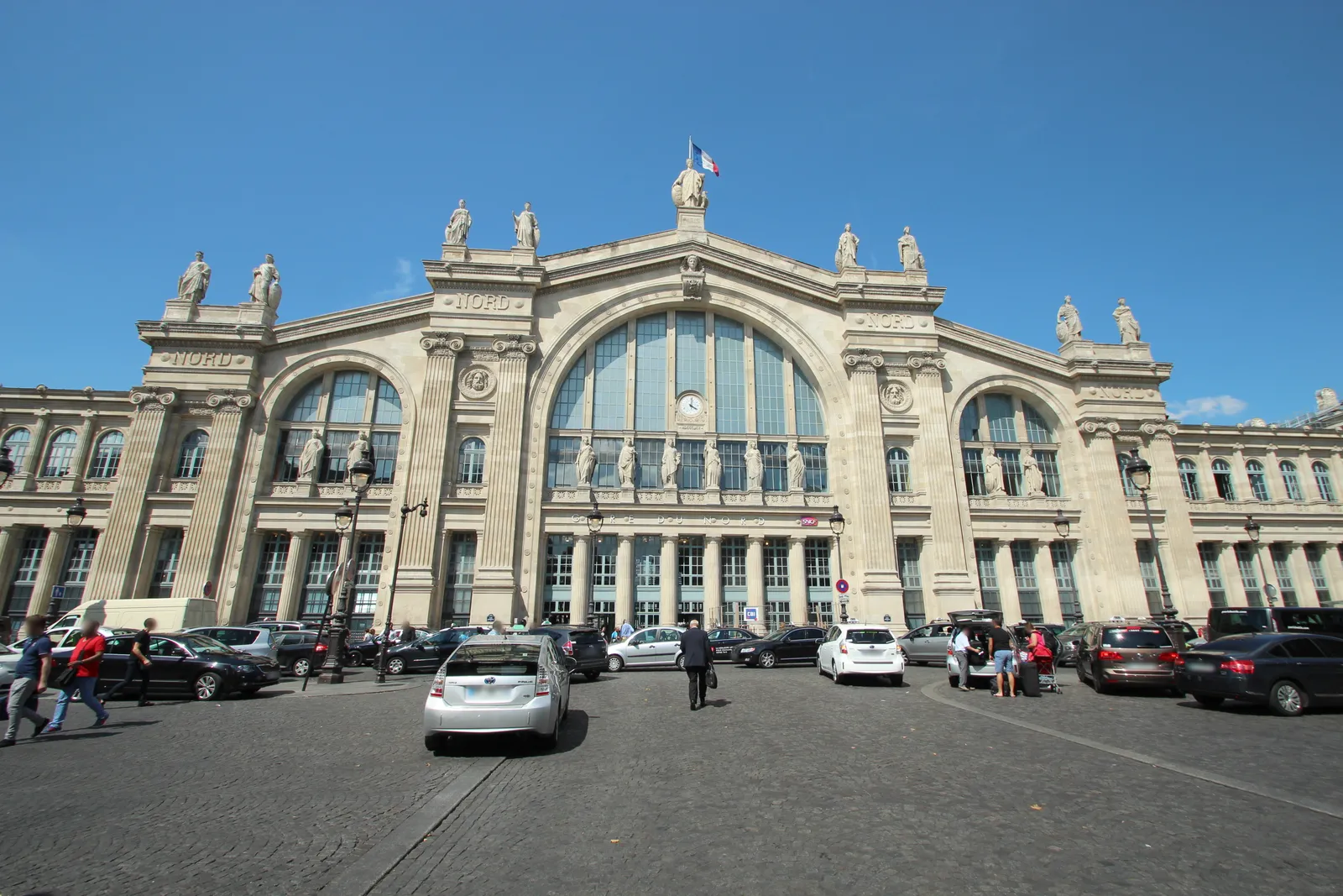 The 1864 front of Paris Gare du Nord, a row of tall stone arches under a line of standing statues along the roofline, with taxis and pedestrians on the forecourt