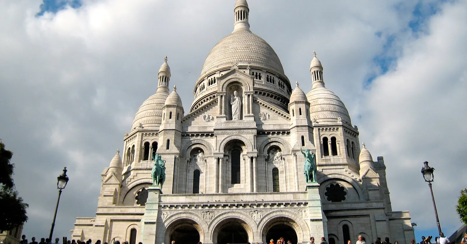 The white stone domes and towers of the Sacré-Cœur basilica in Montmartre, Paris, seen from the steps below with visitors sitting across the terrace in front of it