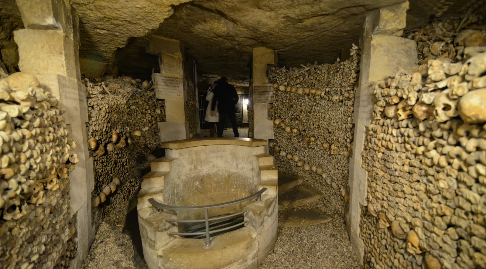 A tunnel in the Paris Catacombs lined on both sides with walls of stacked leg bones and skulls, running away into the lit distance