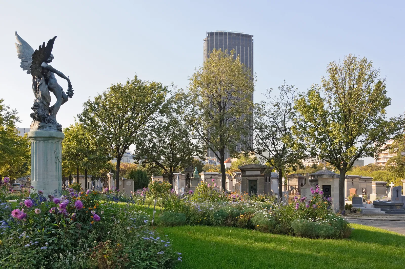 An avenue of tombs in Montparnasse Cemetery in Paris with a winged bronze figure on a plinth at one side, flowerbeds along the path and the Tour Montparnasse standing above the trees