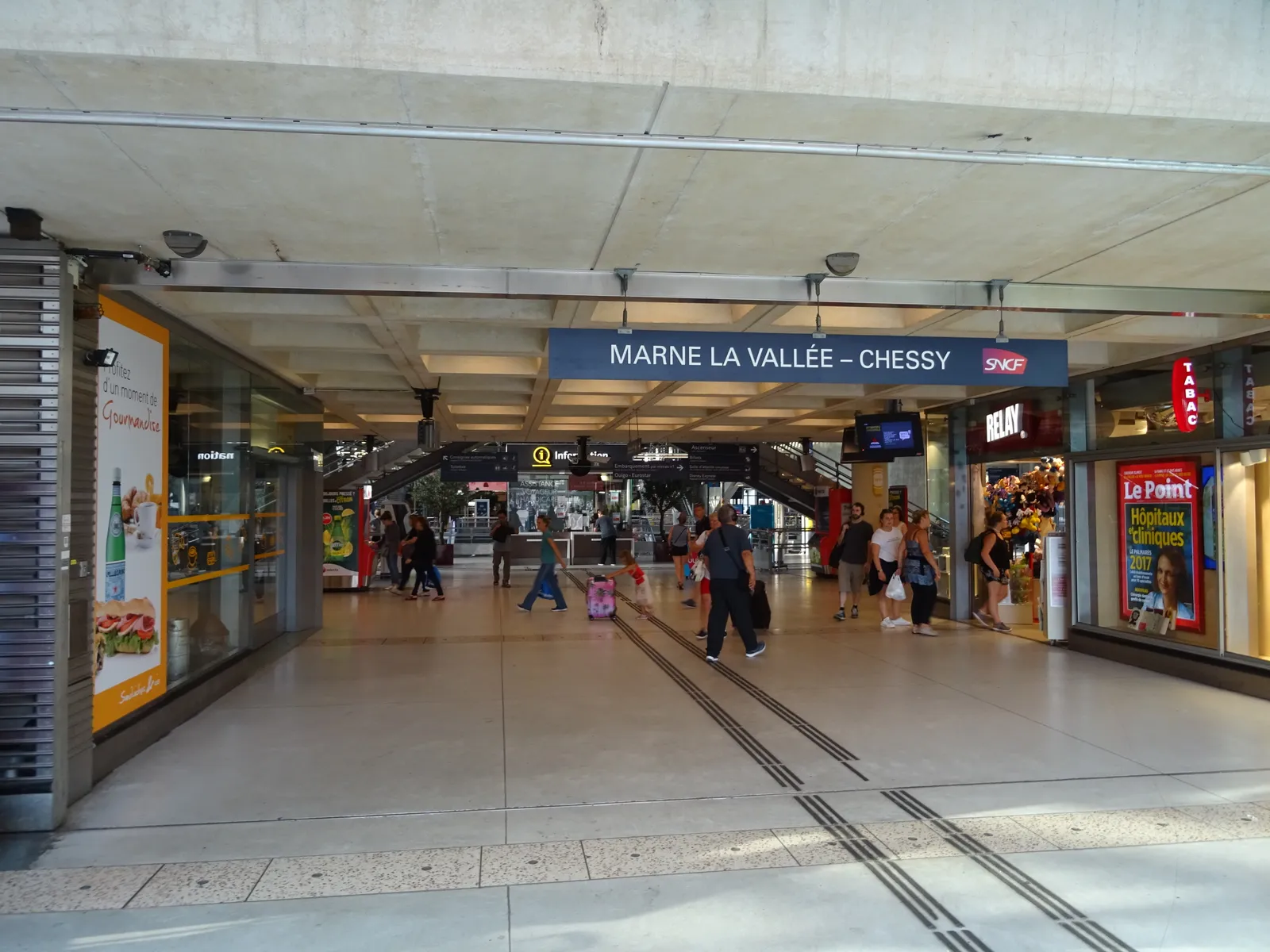 The concourse inside Marne-la-Vallée-Chessy station, a wide passage of shops under a low concrete ceiling with a blue sign reading Marne la Vallée - Chessy hanging above the walkway