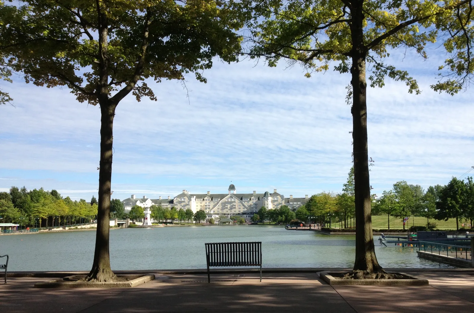 The paved promenade along Lake Disney on a summer morning, a bench between two plane trees and the long white facade of a lakeside hotel on the opposite bank