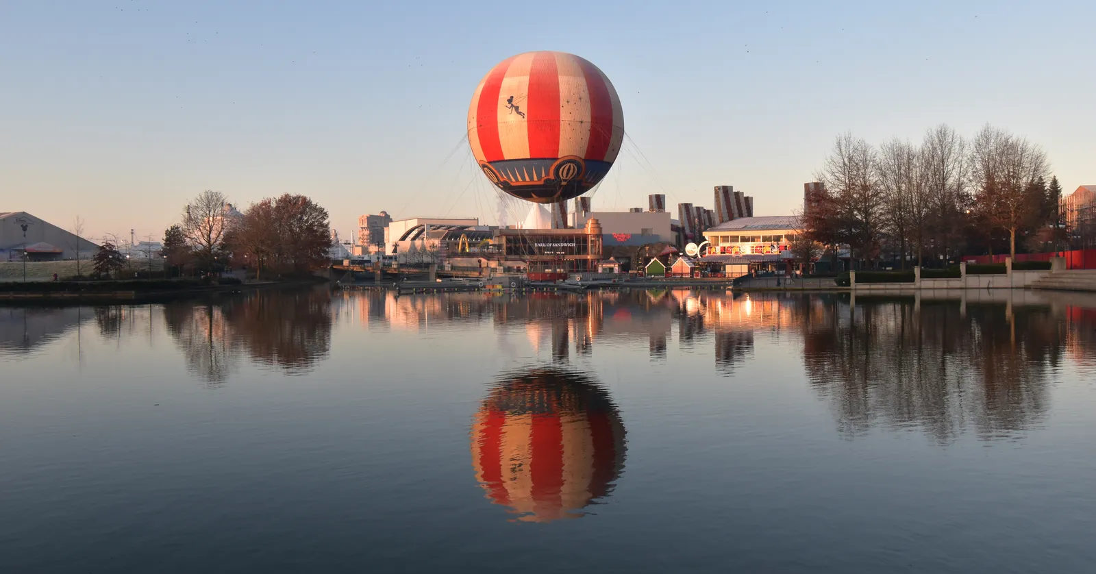 Lake Disney at Disneyland Paris at dusk, a striped tethered balloon hanging above the far shore and mirrored in the still water, with the low roofs of Disney Village behind it
