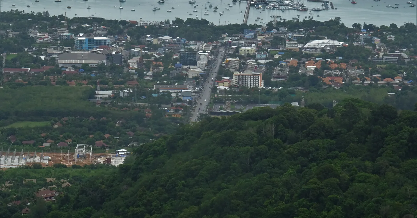 Chalong Bay in Phuket seen from the hills above, the low town spread between forest and shore with a long jetty running out into water crowded with moored yachts and small boats