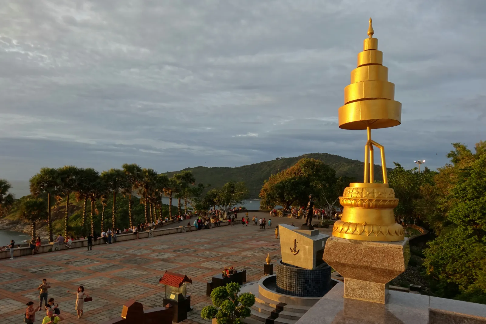 The paved terrace at Promthep Cape in Phuket in late afternoon light, a tall gilded finial in the foreground, palms along the edge and visitors looking out over the headland to the sea
