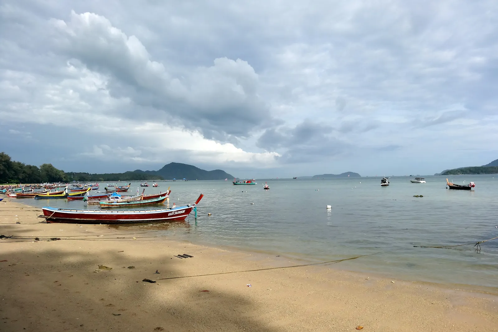 Rawai Beach in Phuket, painted long-tail boats drawn up on wet gold sand under a heavy grey sky, more boats moored offshore and low islands on the horizon