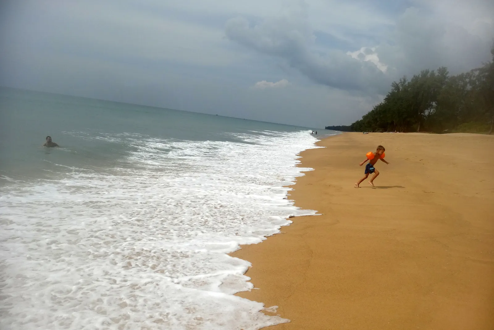 Mai Khao Beach in Phuket under a grey sky, an empty strip of gold sand curving into the distance with white surf breaking along it and a wall of casuarina trees behind