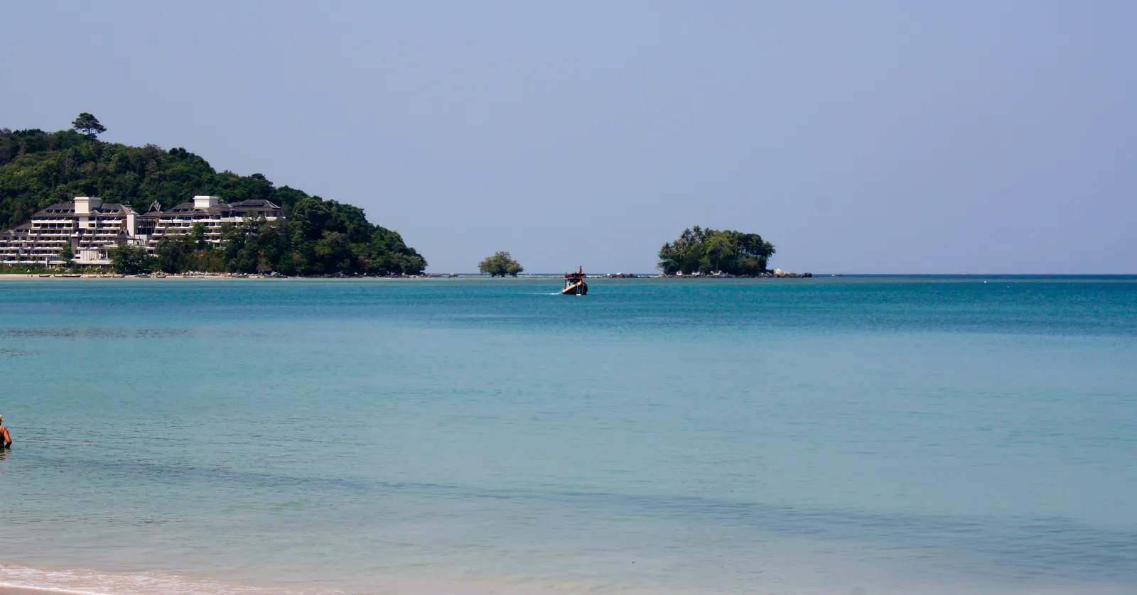 Nai Yang Beach in Phuket, pale turquoise water running up onto white sand with a wooded headland on the left, a small forested islet offshore and a long-tail boat anchored between them