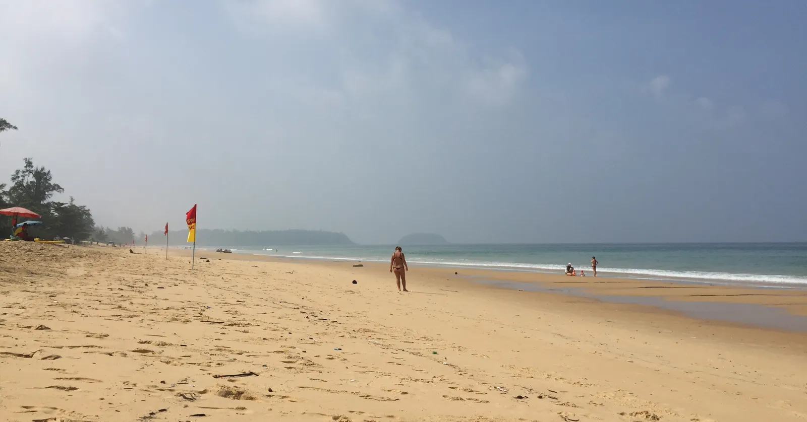 Karon Beach in Phuket, a wide expanse of pale gold sand running into the distance with a red and yellow lifeguard flag planted in it, low surf on the right and a hazy headland at the far end