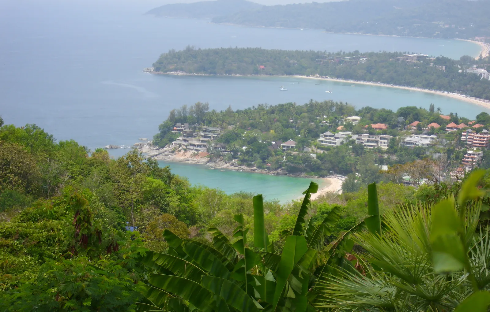 The view from Khao Saam Haad in Phuket, three curved bays with pale sand and turquoise shallows separated by forested headlands, resort rooftops along the far shore and open sea beyond