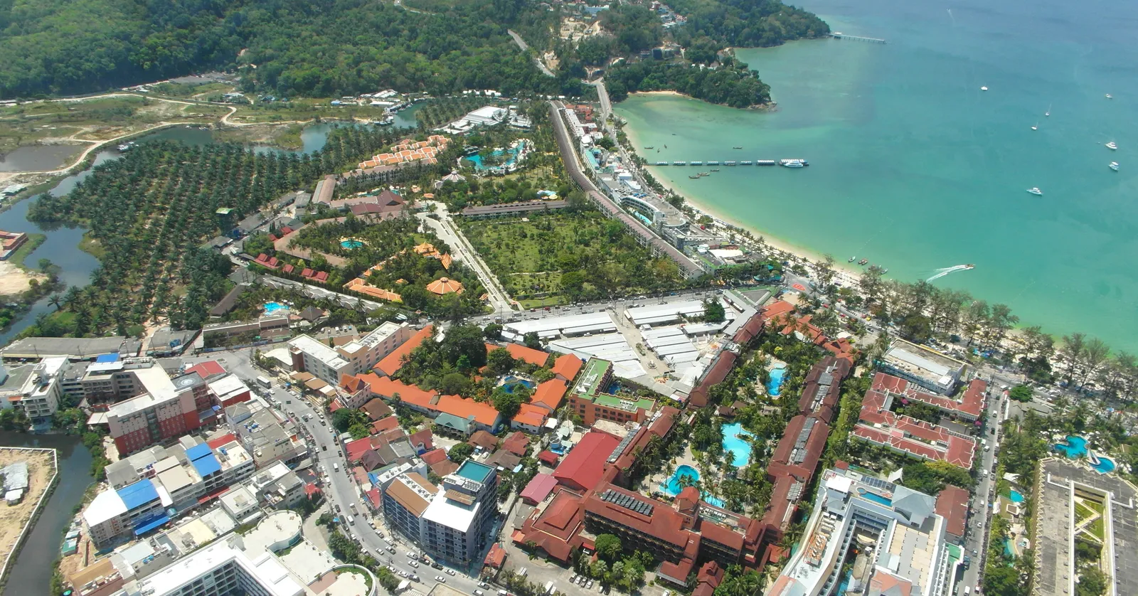 Patong Bay in Phuket from the air, a dense grid of hotels and red-roofed buildings behind a long curve of sand, turquoise water with boats moored offshore and green headlands closing the bay at both ends