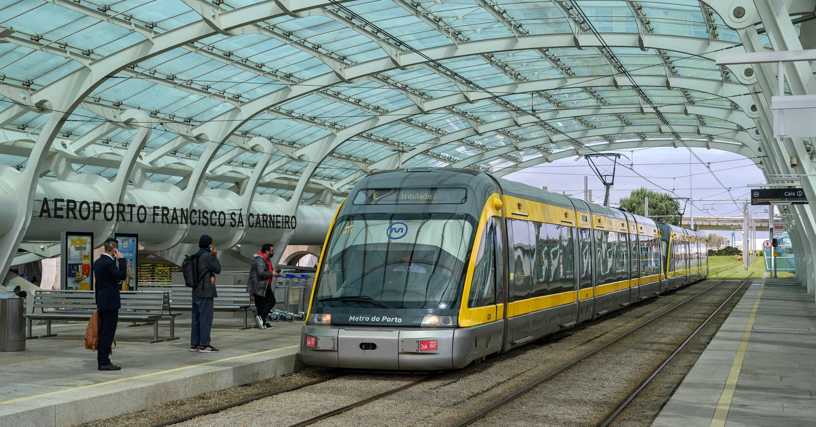 A yellow and grey line E metro train for Trindade waiting under the curved glass roof of Aeroporto station at Porto's Francisco Sá Carneiro airport