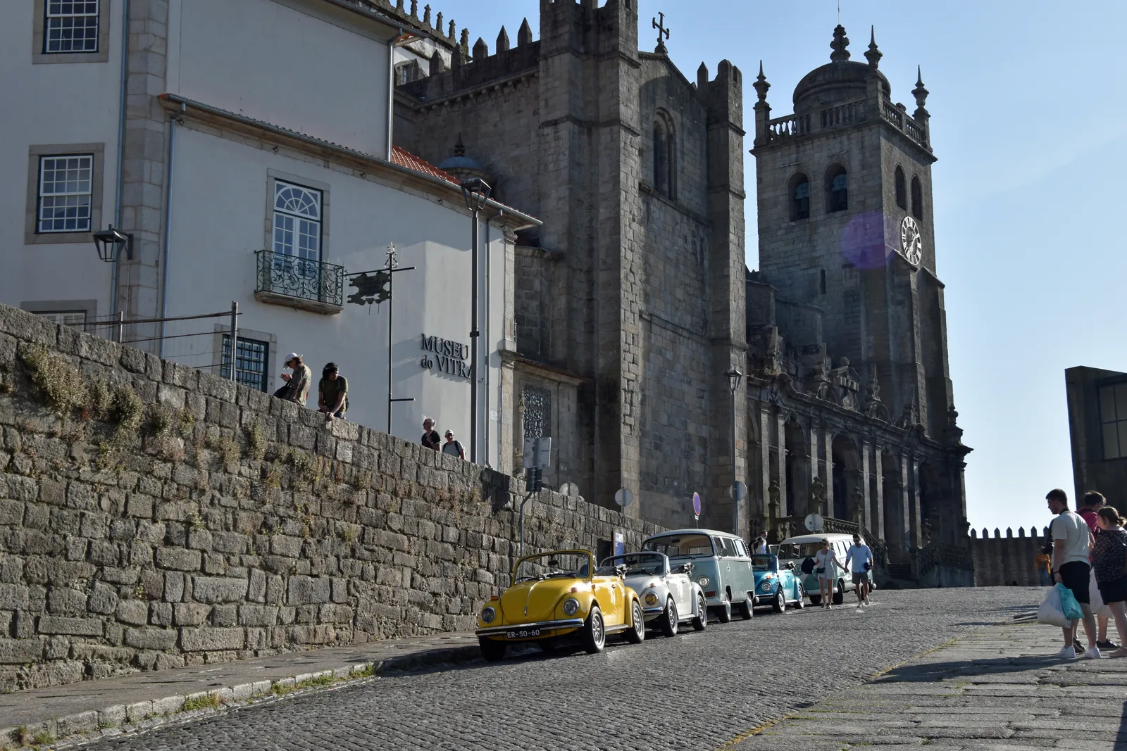 The fortress-like granite towers of Porto Cathedral seen from the cobbled approach below, with vintage cars parked along the terrace wall