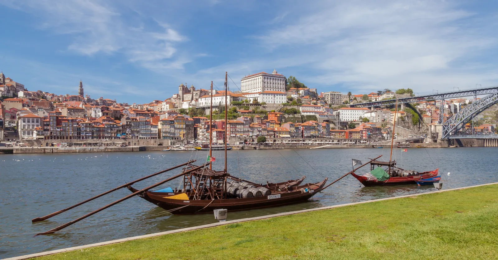 Traditional flat-bottomed rabelo boats moored on the Vila Nova de Gaia bank of the Douro, with Porto's stacked houses and the Dom Luís I bridge across the water